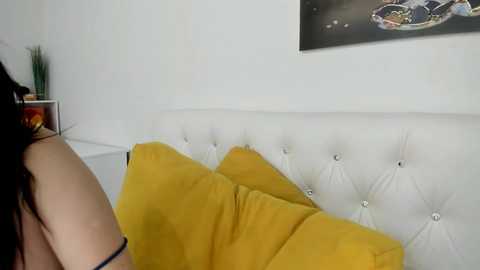 Media: Video of a woman with long black hair, wearing a black bra, sitting on a white tufted sofa with mustard-yellow pillows. The background features a white wall and a framed abstract art piece.