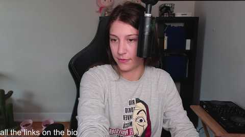 Media: A young woman with shoulder-length dark hair sits in a black gaming chair, wearing a gray T-shirt with a graphic design, in a simple room with a wooden desk, bookshelf, and plants.