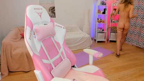 Media: Video of a pink and white gaming chair in a brightly lit bedroom with a woman partially visible in the background, nude, standing in front of a bookshelf.