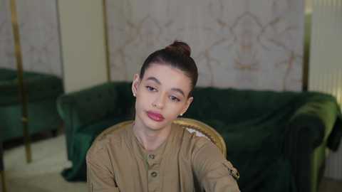 Media: Video of a young girl with light skin, dark hair tied in a bun, wearing a tan military-style jacket, seated in a plush green armchair against a wallpapered backdrop with floral designs.