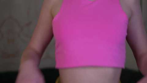 Media: Video of a woman in a bright pink crop top, showcasing her fit physique. The background is blurry, featuring a wall with a faint, indistinct pattern.