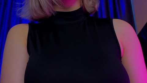 Media: Video of a woman with shoulder-length blonde hair, wearing a sleeveless black turtleneck dress, against a dark blue curtain backdrop with pink and purple lighting.