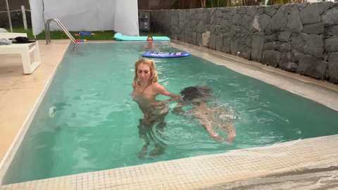 Media: Video of a nude blonde woman with medium breasts standing in a pool, with a partially submerged man behind her. Poolside in a backyard setting, featuring a stone wall and lounge chairs.