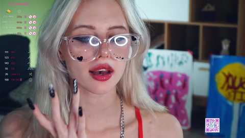 Media: Video of a young, fair-skinned, platinum-blonde woman with large, clear-framed glasses and black-painted nails, wearing a red top, holding a phone, in a colorful room.