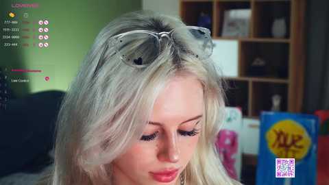 Media: Video of a blonde woman with sunglasses resting on her head, wearing makeup, in a modern living room with a green wall, wooden shelves, and a \"Lucky Brand\" sign.