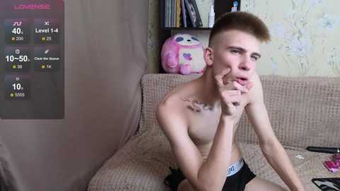 Media: Video of a shirtless, skinny, young man with short hair, wearing black underwear, sitting on a beige couch, holding a phone, in a cluttered room with a pink plush toy and a shelf.