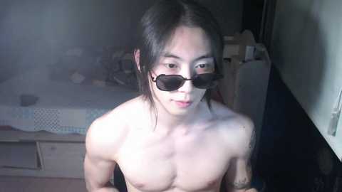 Media: Video of a young, slim, Asian man with long, dark hair, wearing dark sunglasses, sitting topless in a dimly lit room with a cluttered background.