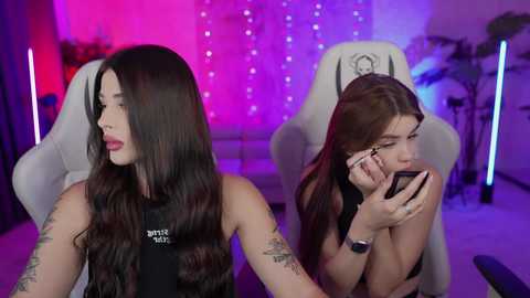 Media: Video of two women with long black hair, one wearing a black tank top, the other in a black bra, sitting in white gaming chairs under purple and pink LED lights, holding smartphones.