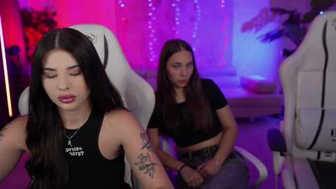 Media: Video of two young women in a gaming room: one with long black hair, wearing a black crop top, and tattoos, the other with long brown hair, in a black crop top, sitting on a white gaming chair.