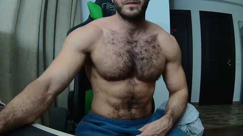 Media: Video of a shirtless, muscular man with dark chest hair, sitting in a green gaming chair. He wears blue shorts, with a towel around his neck. Background includes beige curtains, a door, and a dark wall.