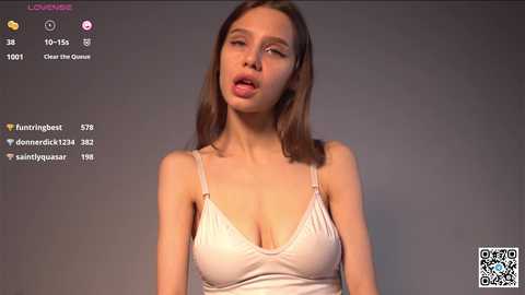 Media: Video of a young Caucasian woman with straight brown hair, wearing a light pink spaghetti-strap tank top, leaning forward with slightly parted lips, against a plain gray background.