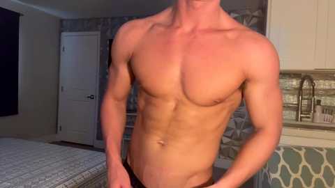 Media: Video of a shirtless, muscular man with fair skin, wearing black underwear, standing in a modern bedroom with patterned wallpaper, white cabinets, and a bed with a grey quilt.