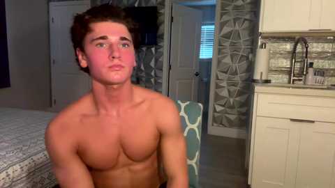 Media: Video of a shirtless, muscular young man with short brown hair and a tan, sitting on a bed in a modern bedroom with a white and geometric-patterned wall, and a kitchenette.