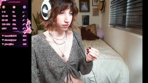 Media: Video of a young woman with shoulder-length wavy brown hair, wearing headphones, a grey cardigan, and a necklace, in a cozy bedroom with a bed, plants, and framed pictures on the wall.