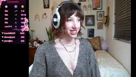 Media: Video of a young woman with light skin and short brown hair, wearing headphones and a V-neck sweater, smiling in a cozy bedroom with wall art, a bed, and streaming controls visible.