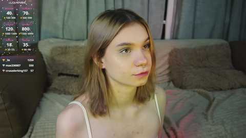 Media: Video of a young Caucasian woman with light skin, straight shoulder-length blonde hair, wearing a white spaghetti-strap top, sitting on a beige sofa with textured cushions. Background includes a grey curtain.