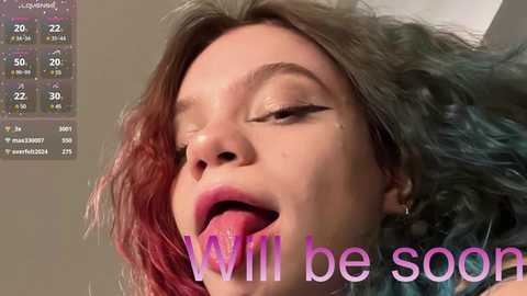 Media: Video of a woman with light skin, long wavy hair dyed pink and teal, wearing black eyeliner and pink lipstick, mouth open, text overlay: \"I will be soon.\