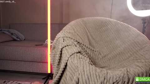 Media: Video of a beige, textured, ribbed chair with a blanket draped over it, next to a modern, minimalist bed with a gray cushion and a red light strip on the left side, in a softly lit room.