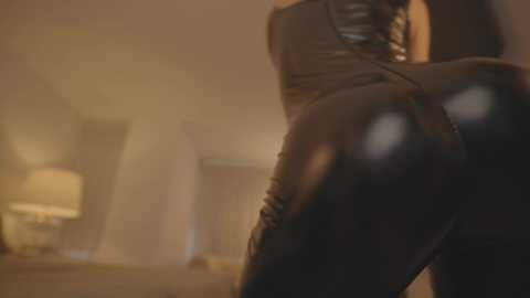 Media: Video of a woman in a tight, black leather bodysuit, emphasizing her curvaceous, round buttocks, captured in a dimly-lit room with soft, blurred background.