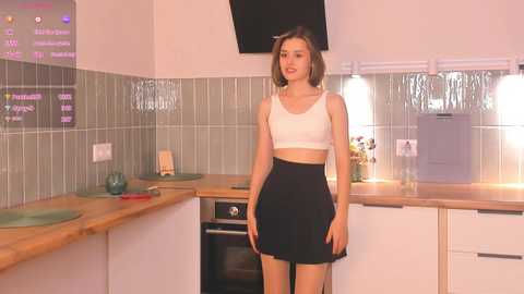 Media: Video of a young woman with a slim build, wearing a white crop top and black skirt, standing in a modern kitchen with green tiled backsplash and wooden countertops.