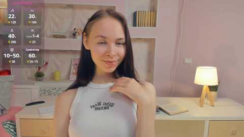 Media: A young Caucasian woman with long black hair, wearing a white tank top with \"These days, I feel like\" printed on it, sits in a cozy, pastel-colored bedroom with a white desk and lamp.