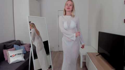 Media: Video of a blonde woman in a white, sheer, long-sleeved dress, posing in a modern, minimalist living room with light wood floors and a gray sofa.