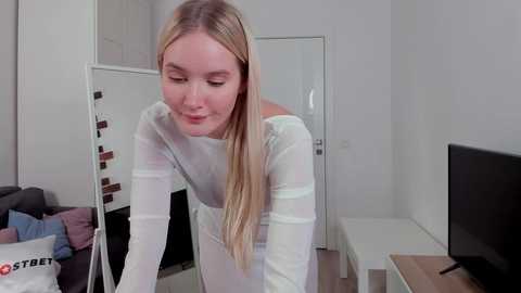 Media: Video of a blonde woman in a white long-sleeve top, leaning over a mirror in a modern, minimalistic living room with a flat-screen TV on a white stand.