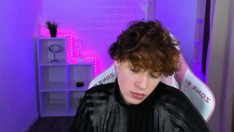 Media: Video of a young person with curly brown hair, wearing a black cape, seated in a white chair with purple LED lights behind them.