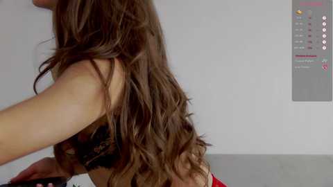 Media: Video of a light-skinned woman with long, wavy brown hair, wearing a black lace bra, captured from the side. Background includes a white wall with a red and white calendar and a gray surface.