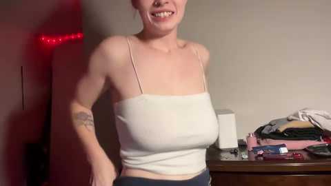 Media: Video of a smiling, slender, fair-skinned woman with short hair, wearing a white spaghetti-strap crop top and dark pants, in a dimly lit room with a red string of lights, cluttered table, and beige wall.