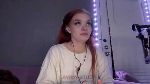 Media: Video of a young woman with long, straight red hair, wearing a white t-shirt, black headphones, and a neutral expression, in a dimly lit room with purple LED lights and a camera on a tripod.