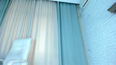 Media: Video of a minimalist room featuring a white leather chair, beige and light blue curtains, and a white brick wall with an air conditioning unit on the right.