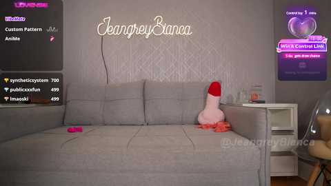 Media: Video of a modern living room with a gray sectional sofa featuring a large, pink, dildo-shaped plush toy. Textures include soft upholstery, glass shelves, and a digital TV screen displaying streaming content.