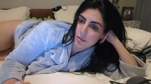 Media: Video of a woman with long black hair, light skin, and medium breasts, lying on a bed in a light blue robe, in a softly lit bedroom with beige walls and white pillows.