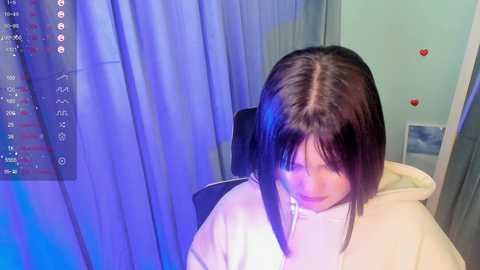 Media: Video of a young Asian woman with long, straight black hair, wearing a white top, seated in a dimly lit room with blue curtains and heart stickers.