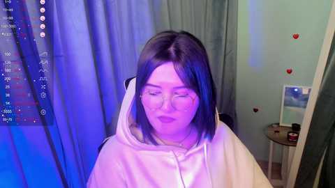 Media: Video of a young woman with long black hair, wearing glasses and a white hoodie, standing in a room with blue curtains, heart stickers on the wall, and a mirror.