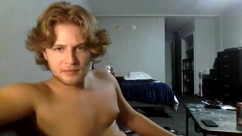 Media: A video of a shirtless, fair-skinned man with shoulder-length, wavy brown hair, sitting at a cluttered desk in a dimly lit room with a blue bed and open closet door.