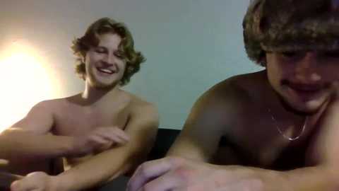 Media: Video of two shirtless young men, one with curly hair, grinning, the other with a mustache, lying on a couch, dimly lit room.