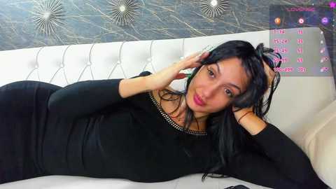 Media: Video of a young woman with long black hair, lying on a white tufted couch, wearing a black dress. Background features a textured, metallic wall with circular patterns and a digital clock displaying time and date.