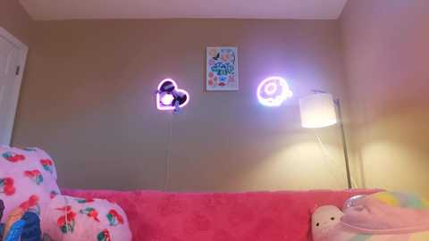 Media: A video of a cozy, pink-fur-covered sofa in a child's bedroom, featuring a stuffed toy, colorful blankets, and glowing neon spiral lights on beige walls.