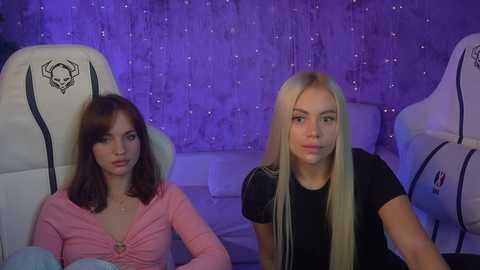 Media: Video of two young women with long hair in a dimly lit room with purple lighting. One wears a pink top, the other a black top.