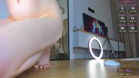 Media: Video of a nude woman with a large breast, crouching on a wooden floor, in a modern room with a TV and a ring light.