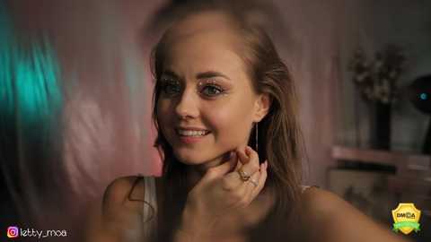 Media: Video of a young woman with fair skin, long brown hair, and green eyes, smiling, wearing a sleeveless top, in a dimly lit room with blurred background.