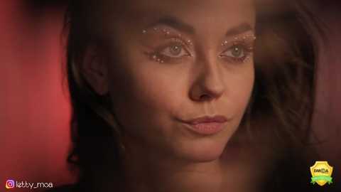 Media: Video of a close-up of a woman with light skin, dark hair, and makeup including glittery eyeshadow. Background is blurred red and brown, suggesting a warm, cozy setting.