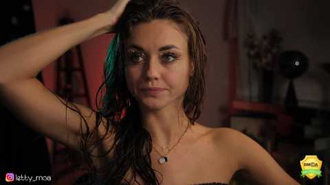 Media: Video of a young woman with wet, dark hair, wearing a strapless top, standing indoors. She has a confident expression. Background includes a dimly lit room with furniture and plants.