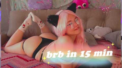 Media: Video of a young, curvy woman with long, pastel pink hair, wearing cat ears, glasses, and black lingerie, lying on a red-patterned blanket in a cozy bedroom with plush toys and fairy lights. Text reads \"bri in 15 min.\