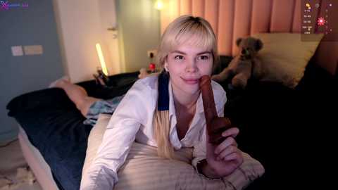 Media: Video of a blonde woman with a small penis in her mouth, lying on a bed in a dimly-lit room with a teddy bear, pink curtains, and a glowing lamp.