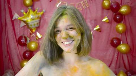 Media: Video of a smiling, topless young woman with shoulder-length blonde hair, covered in bright yellow and orange Holi powder, against a red backdrop adorned with golden balloons, a crown, and banners.