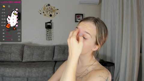 Media: Video of a fair-skinned woman with light brown hair, wearing a necklace, covering her eyes with her hand, sitting on a gray couch in a living room.