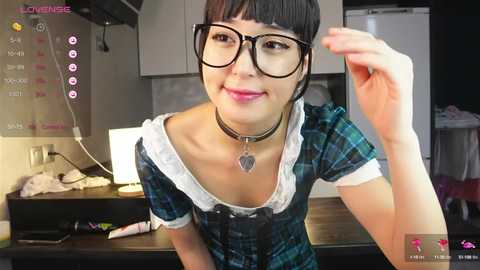 Media: Video of an East Asian woman with short black hair, wearing black-rimmed glasses, a green plaid dress, and a choker necklace, smiling in a modern kitchen with a white fridge and black counter.